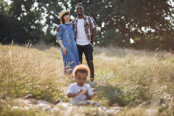 Blur foreground of little african boy sitting on grass and playing while his multiracial parents standing behind, hugging, smiling and looking at him. Happiness from parenthood.