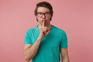 Studio shot of young handsome male, wears glasses and blue t-shirt, looks into camera and show silence gesture with serious facial expression. Isolated over pink background