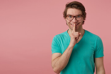 Studio shot of young handsome male, wears glasses and blue t-shirt, looks into camera and show silence gesture with serious facial expression. Isolated over pink background