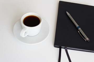 The concept of a workplace with coffee in a neat white mug on a saucer and a notebook with a pen on a modern white office desk. Break for a delicious and hot coffee. Easy to use. Minimalism. Side view