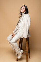 full length of cheerful woman in suit posing while sitting on wooden stool on beige