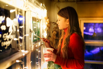 Cute caucasian child girl in a red dress paints the windows.Kid girl decorates the house for christmas, paints the windows, dark style and festive lights on the background