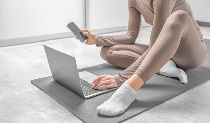 Fototapeta premium Girl in sportwear sitting on the training mat and searching for online training on the laptop and phone.