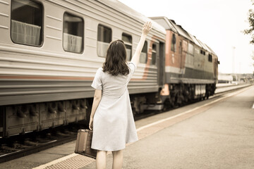 Young woman in with a suitcase waves to the departing train. Retro-style processing. The concept of being late for a train and seeing off or meeting a young person from a train on the platform.