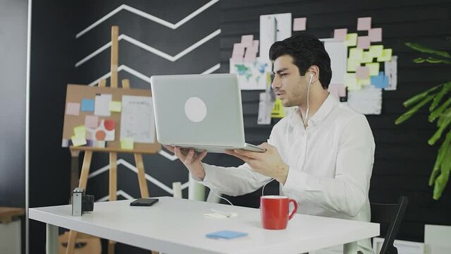 A young man of Caucasian appearance is sitting at a computer and working remotely. A young man wearing headphones communicates via video link and shows his notes on the wall