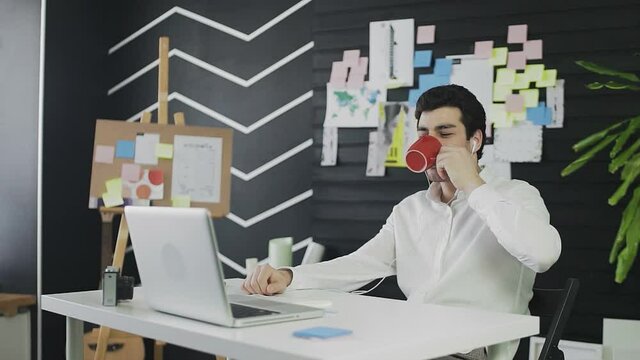 A young man of Caucasian appearance is sitting at a computer and working remotely. A young man wearing headphones communicates via video link and drinks tea or coffee
