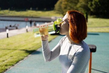 fit ginger woman in sport clothes drinking water while working out on sports ground in autumn sunny...