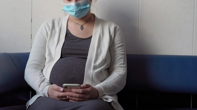 Young Pregnant Woman In Medicine Protective Mask Reading On Couch Waiting For Appointment Medical Control On Hospital. Visit Gynecologist Doctor At Clinic. Examine Expectant Belly Healthcare Check Up