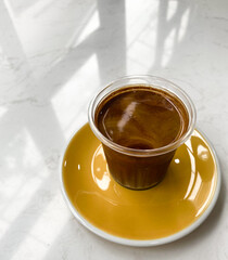 Milk coffee in a clear plastic cup rests on a yellow saucer set on a white marble table. and the shadow of the window reflected down