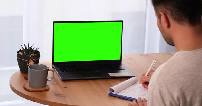Man taking notes with personal computer mock-up chroma key display, isolated green screen PC for easy replacement. The Caucasian guy at home in bright living room