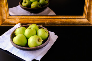 Green apples in an old clay plate on a white napkin on a black background. Reflection in the mirror. Beautiful antique mirror in a gold frame. Empty space for your text and design. Culinary template. 
