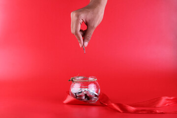 A human hand holds a coin and will put it in a jar decorated with a red ribbon. red background. 