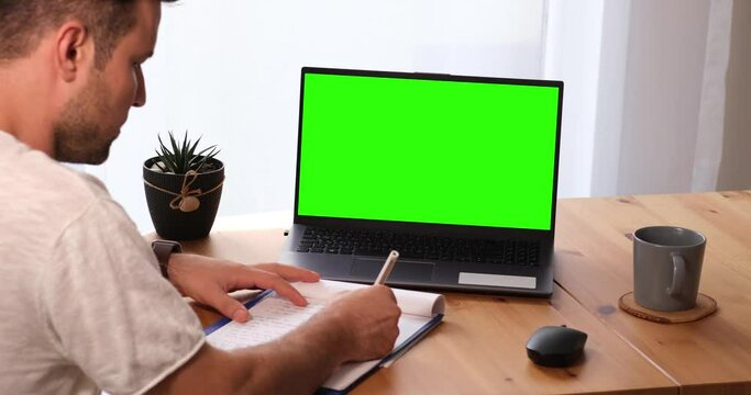 Man taking notes with personal computer mock-up chroma key display, isolated green screen PC for easy replacement. The Caucasian guy at home in bright living room