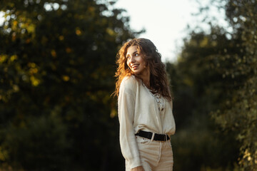 Young beautiful happy woman with a cute smile and curly hair with a knitted sweater walks in the park on a sunny summer day. Pretty female smiling face