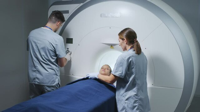 Slowmo Shot Of 9-year-old Girl Lying Down On Scanner Table Before Doing Examination In Care Of Two Radiologists Operating Machine Equipment At Modern Clinic