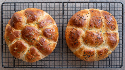 Two traditional home cooked Jewish Challah bread loaves, on a wire tray. One is topped with nigella seeds and the other with rosemary. 