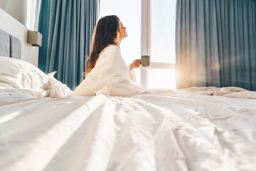Pretty positive woman with long hair drinks water and sitting at bed in sunny lazy morning.