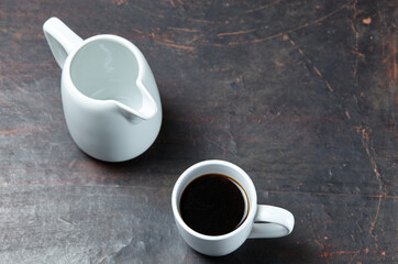 White cup with coffee and empty milk jug on a white wooden background, closeup. Energy breakfast, morning routine concept, top view