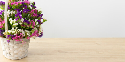 A basket with multicolored statice flowers stands on a wooden table top on a light background . The flowers are purple, white and pink . A banner with free space for your business .