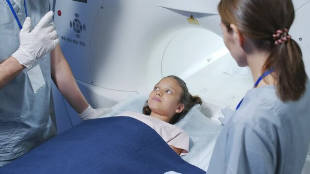 Slowmo shot of male doctor and his assistant explaining stages of MRI procedure to little girl lying on scanner table in radiology room of modern clinic