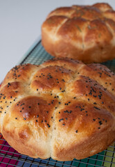 Two traditional home cooked Jewish Challah bread loaves, on a wire tray. One is topped with nigella seeds and the other with rosemary. 