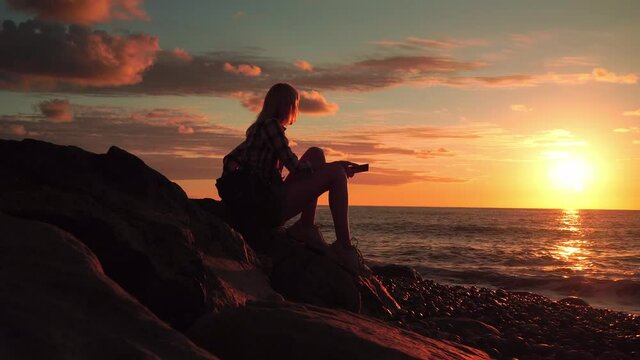 silhouette of slender woman on beach at beautiful sunset, she is sitting dreaming, she has mobile phone or smartphone in her hands. Red scarlet sunset onclouds sky. waves are beating against shore