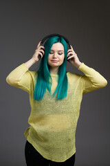 Obraz na płótnie Canvas Girl with headphones listens to music or podcast. Woman with blue green hair in a yellow sweater isolated on a blue background. Bright colored look
