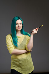 The girl artist holds a brush with paint. Woman with blue green hair in a yellow sweater isolated on a grey background. Bright colored appearance of a creative person