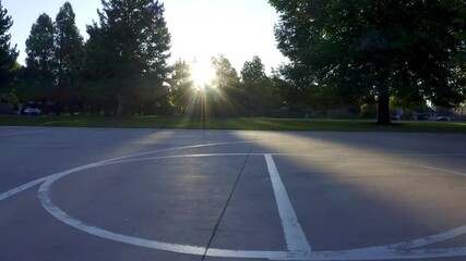 Low angle tracking shot over basketball court in an urban American park, looking into sun, with lens flare and long shadows, in morning light through oak trees, captured by a drone in 4K resolution. - Powered by Adobe