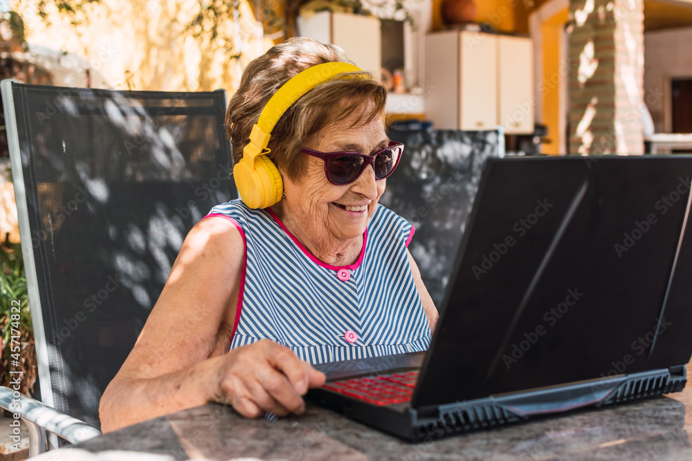 Wall mural old woman smiling with headphones and sunglasses making a video call outdoors