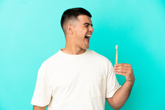 Young handsome man brushing teeth over isolated blue background laughing in lateral position