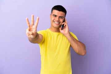 Young handsome man using mobile phone over isolated purple background happy and counting three with fingers