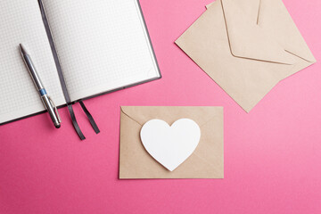 The concept of a romantic love letter for Valentine's Day. A closed envelope made of kraft paper with a white heart on a pink background and an open notebook with a pen. A declaration of love. Closeup