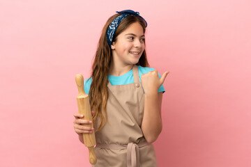 Little caucasian girl holding a rolling pin isolated on pink background pointing to the side to present a product