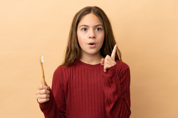 Little caucasian girl brushing her teeth isolated on beige background thinking an idea pointing the...