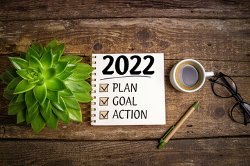 New year goals 2022 on desk. 2022 resolutions list with notebook, coffee cup and eyeglasses on wooden background. Goals, plan, strategy, business, idea, action concept. Top view
