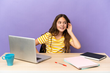 Little student girl in a workplace with a laptop isolated on purple background listening to something by putting hand on the ear