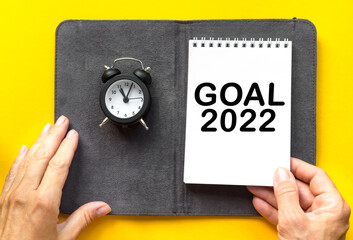 GOAL 2022 women's hand holds NOTEPAD AND a small alarm clock yellow background