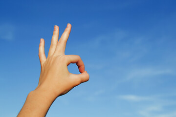 A hand against a blue sky shows an okay gesture, performed by connecting the thumb and forefinger and keeping the other fingers straight or relaxed from the palm, copying space.