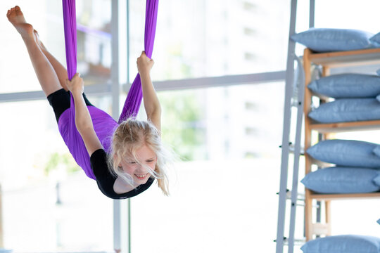  young smiling girl practice in stretching swing in purple hammock in fitness club. kids Aerial flying yoga exercises. - Powered by Adobe