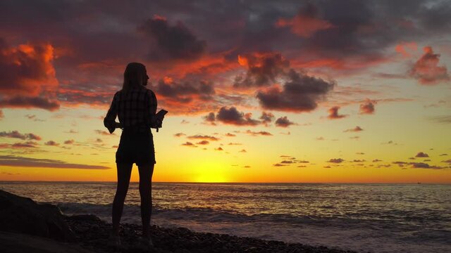 silhouette of slender woman on beach at beautiful sunset, she stands and dreams, takes photo on her phone. she has smartphone in her hands. red scarlet sunset on cloudy sky. waves are beating on shore
