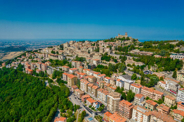 Fototapeta na wymiar Aerial view of San Marino old town with old buildings and red roofs on the hill on a sunny day with clear sky. Picture from above with Italy behind in horizon