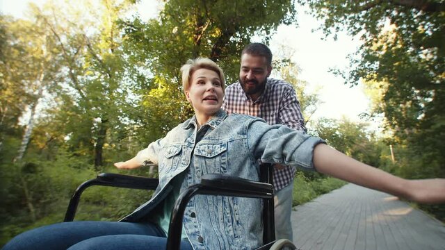 Happy man rolls emotional disabled woman in wheelchair, who spread her arms like wings, through park, side down view. Concept of self-acceptance and support for people with disabilities.