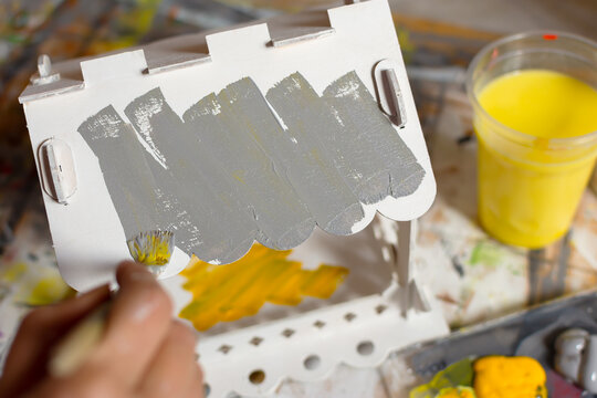 Wooden bird feeder painted in gray and yellow colors. Trending shades 2021. Creative homework with children. A brush in the hands of a close-up. Artistic process with acrylic paints