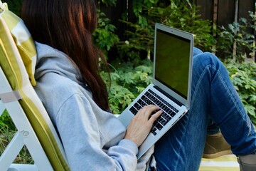 Young lady with red hair working on a silver laptop with green screen wearing grey hoodie and jeans and sitting in a chaise lounge in a green garden. Freelancer freelance job work from home concept