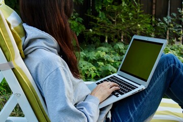 Young lady with red hair working on a silver laptop with green screen wearing grey hoodie and jeans and sitting in a chaise lounge in a green garden. Freelancer freelance job work from home concept