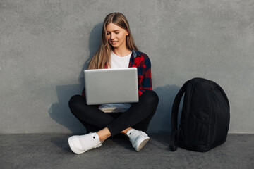 Young woman freelancer, with a backpack, sits on the street at free time with a new modern laptop, using a laptop in the city near the gray city wall