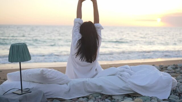 Back view of slim young woman stretching sitting on white cushioned bed at sunrise on sea coast. Happy relaxed Caucasian brunette lady waking up on sandy beach admiring beauty of nature. Slow motion