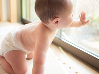ハイハイしながら窓に手をつき外を見つめるおむつをした赤ちゃん