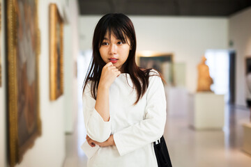 Thoughtful chinese female visitor with guide-book looking at artwork painting in the museum indoors